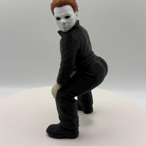 May include: A small figurine of a character with a white mask, short brown hair, and a black jumpsuit. The figure is posed in a crouching position with its back arched, wearing black boots and showing light-colored hands resting on its knees.