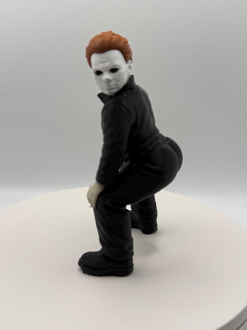 May include: A small figurine of a character with a white mask, short brown hair, and a black jumpsuit. The figure is posed in a crouching position with its back arched, wearing black boots and showing light-colored hands resting on its knees.