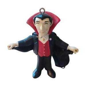 Puede incluir: Una figura de vampiro con rostro pálido, colmillos visibles, cabello negro y una capa negra forrada en rojo. El personaje viste una chaqueta negra sobre un chaleco rojo con botones dorados, camisa blanca y pantalones negros, con una argolla metálica.