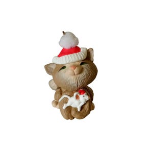 May include: A tan cat figurine wearing a red and white Santa hat and holding a white mouse with a Santa hat. The cat has green eyes and a smiling expression. A small gold loop is at the top for hanging.