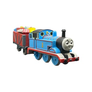 May include: A blue toy steam locomotive with the number 1 printed in yellow on its side, featuring a grey face, black chimney, and red accents. Attached to the back is a red cargo wagon filled with various colorful, indistinct small items.