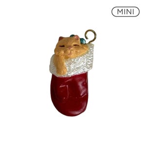 May include: A miniature ornament featuring a light orange cat with closed eyes resting inside a red stocking with a white textured cuff. The ornament includes a gold-colored metal hook for hanging and a small label with the text MINI.