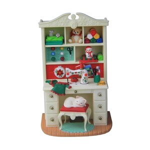 May include: A miniature cream-coloured sewing desk hutch featuring shelves stocked with yarn, a teddy bear, and buttons. The desk includes a white sewing machine, a red pincushion, a green garment, and a white cat resting on a red-topped stool.