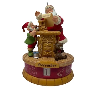 May include: A decorative Christmas calendar figurine featuring Santa Claus and an elf at a workbench. Santa holds a scroll labeled Michele, while the elf paints a toy train. A teddy bear sits nearby. The base displays the word December and the number 1.