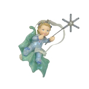 May include: A decorative figurine of a baby with blonde hair wearing a light blue outfit with glittery accents. The baby is positioned on a green leaf-like base and holds a silver wand topped with a star-shaped arrangement of clear beads.