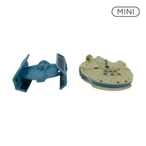 Puede incluir: Dos adornos en miniatura de naves espaciales con la etiqueta MINI. Uno es un caza TIE azul con paneles de ala negros, y el otro es un Halcón Milenario circular de color beige con detalles grises y acentos rojos. Ambos tienen pequeñas anillas.