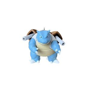 May include: A blue and tan creature figurine with a rounded shell on its back and two cylindrical cannons extending from the sides. The figure has a light blue head, a tan chest plate, and white claws on its feet and hands, featuring a small metal loop.