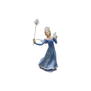 May include: A figurine of a fairy wearing a long blue dress with white snowflake patterns and a white lace hem. The fairy has translucent wings, a white crown, and holds a thin wand topped with a white star in the right hand.