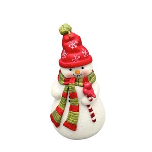 May include: A white snowman figurine wearing a red knit-style hat with white snowflake patterns and a green and red striped scarf. The snowman has a small orange carrot nose, black dot eyes, and holds a red bird perched on a candy cane.
