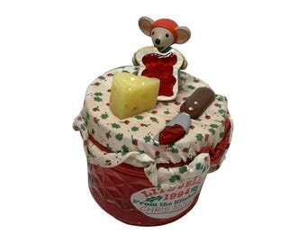 May include: A red glass jar with a fabric lid cover featuring a holly pattern. A mouse figurine wearing an orange hat holds a piece of toast with red jam. A yellow cheese wedge and a small brown-handled knife with red jam sit on the lid.