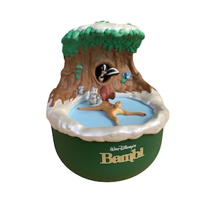 Op de afbeelding: Een ronde groene decoratieve voet bevat de tekst Walt Disney's Bambi in goud. Bovenop toont een gebeeldhouwd tafereel een bruin reekalfje dat op een lichtblauw oppervlak ligt, omringd door een kleine grijze eekhoorn, een witte muis en een stinkdier.
