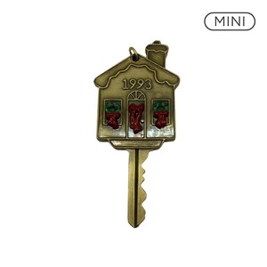 Könnte beinhalten: Ein messingfarbenes Metallschlüssel-Ornament in Form eines Hauses mit Schornstein, auf dessen Vorderseite die Jahreszahl 1993 eingeprägt ist. Das Hausdesign zeigt zwei Fenster mit grünen Kränzen und eine mittlere Tür mit roter Schleife. Das Wort MINI steht darüber.