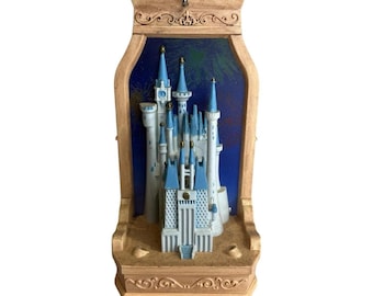 Peut inclure: Un présentoir mural décoratif présentant un modèle tridimensionnel de château aux murs blancs et aux toits coniques bleus. Le château est placé sur un fond bleu foncé avec des motifs de feux d'artifice, le tout encadré par une structure beige.