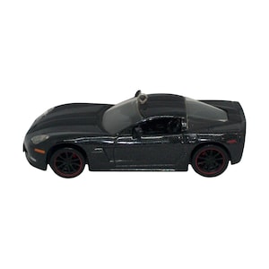 May include: A black sports car model with a metallic finish, featuring dark wheels with thin red rims, clear windows, and a small silver metal loop attached to the roof for hanging. The vehicle has a low-profile design with visible headlights.