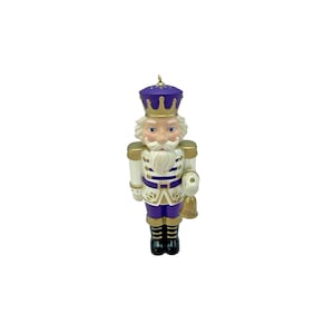 May include: A nutcracker ornament featuring a purple and white uniform with gold accents. The figure wears a purple crown-shaped hat, has a white beard, and holds a small gold bell in one hand. The bell is clearly inscribed with the year 2012.