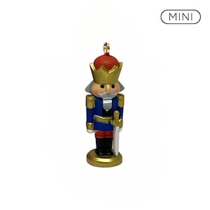 May include: A miniature nutcracker ornament featuring a red hat, gold crown, blue jacket, and black boots. The figure holds a silver sword and stands on a gold base. The text MINI is displayed in a rounded rectangular border above the figure.