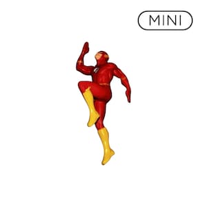 May include: A figurine of the superhero The Flash in a dynamic running pose, wearing a red suit with yellow boots and a lightning bolt emblem on the chest. The figure is positioned below a white oval badge containing the black text MINI.