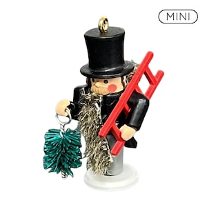 May include: A mini nutcracker ornament wearing a black top hat and coat, featuring a textured gold beard, pink cheeks, and a red ladder. The figure holds a metallic green chimney brush in one hand and has a gold hanging loop at the top.
