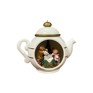 Könnte beinhalten: Ein weißes, teekannenförmiges Ornament mit einem hohlen Zentrum, das eine Miniaturszene mit einem Teddybären und einer Figur an einem Tisch mit Teetassen zeigt. Die Teekanne hat goldfarbene Verzierungen, einen Henkel und einen Ausguss.