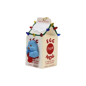 Puede incluir: Un adorno en forma de cartón de ponche de huevo color crema con una figura de pájaro azul en el frente. El cartón está decorado con una cadena de luces navideñas multicolores y el texto "EGG NOG", "ENJOY BY 12-25-92" y "VITAMINS AND CHEER ADDED".