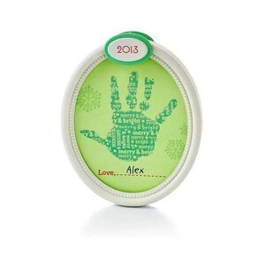 May include: An oval-shaped white frame containing a light green insert with a dark green handprint graphic. The handprint is filled with the repeating text "merry & bright". The top features a green tab with "2013" and the bottom says "Love, Alex".