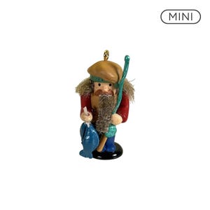 May include: A miniature nutcracker ornament featuring a figure wearing a tan cap, red jacket, and blue pants. The figure has a brown beard, holds a blue fish, and carries a green fishing pole. The text MINI is displayed in a white oval.
