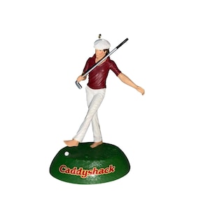 May include: A figurine of a barefoot golfer wearing a white cap, a maroon polo shirt, and white trousers, holding a golf club. The figure stands on a green base with a small white golf ball and the red text Caddyshack printed on the front.