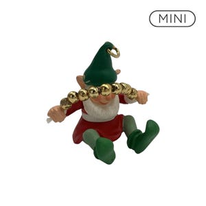 May include: A small elf figurine wearing a green hat, red tunic, and green shoes, holding a string of shiny gold-colored beads across its face. The figure has a white beard and a gold metal loop at the top of the hat. The text MINI is visible.