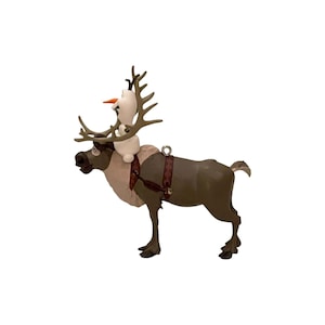 May include: A brown reindeer figurine with a cream-colored chest patch and a dark brown harness, featuring a small white snowman character with an orange carrot nose perched between the reindeer's large antlers. A silver metal loop is attached to the back.