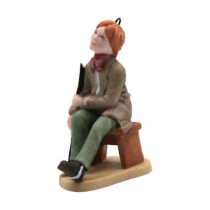 Pode incluir: Uma estatueta de cerâmica de uma pessoa com cabelo ruivo, vestindo uma jaqueta bege, um cachecol vermelho escuro e calças verdes. A figura está sentada num pequeno banco castanho, olhando para cima e segurando um objeto preto ao lado.