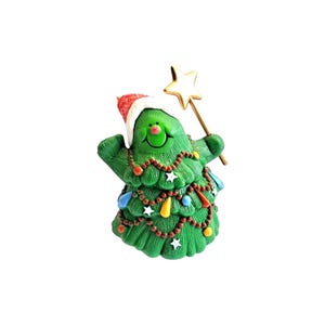 Può includere: Una statuetta a forma di albero di Natale verde con un cappello rosso e bianco. L'albero è decorato con ghirlande di perline rosse, piccole stelle bianche e ornamenti colorati a forma di goccia. Impugna una bacchetta a stella dorata.