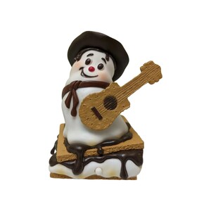 May include: A white snowman figurine wearing a dark brown hat and scarf, holding a light brown acoustic guitar. The snowman is positioned on top of a s'more, which consists of two light brown graham crackers with white marshmallow and dark brown chocolate.