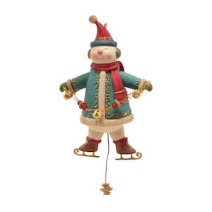 Puede incluir: Un adorno de muñeco de nieve con gorro rojo, abrigo verde azulado con ribetes dorados y bufanda roja. El muñeco tiene orejeras verdes, patines de hielo marrones y sostiene una cuerda con cascabeles dorados y cuentas rojas. Cuelga un hilo blanco.