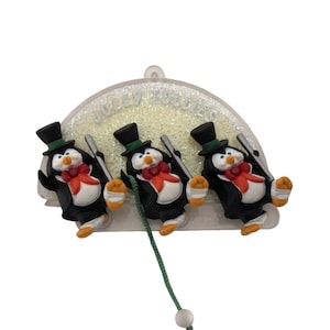 May include: A Christmas ornament featuring three penguin figures in black top hats and tuxedos, each holding a silver cane. They have red bow ties and orange feet. The ornament has the text "JOLLY HOLIDAY" and a green string.