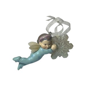 May include: A hanging ornament featuring a small figure with brown hair and a pink headband, wearing a light blue outfit with snowflake patterns. The figure has translucent wings and is holding a large, silver, glittery snowflake decoration.