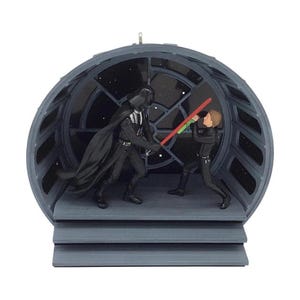 以下が含まれることがあります： 黒いスーツを着た2人のフィギュアがライトセーバーで決闘しているミニチュアシーンの装飾品。一方は黒いヘルメットとマントを着用し、もう一方は緑色のライトセーバーを手にしている。円形のフレーム内の灰色のプラットフォームの上に立っている。