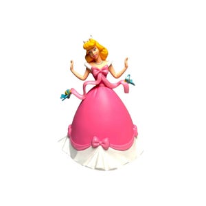 Puede incluir: Una figura de un personaje rubio con un vestido rosa y una falda interior blanca. El vestido tiene un lazo rosa grande en la cintura y un lazo más pequeño en el dobladillo. Dos pájaros azules pequeños se posan sobre las cintas rosas flotantes.