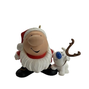 May include: A decorative figurine featuring a round Santa Claus with a red hat, white beard, and black boots standing next to a small white reindeer with a blue nose and brown antlers. The ornament has a gold loop at the top for hanging purposes.