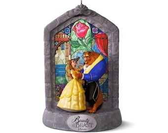 Puede incluir: Una figura decorativa que muestra a Bella y la Bestia bailando frente a un vitral. La base es de material gris similar a la piedra con una placa plateada que dice Beauty and the Beast 25 Years. Una rosa roja está representada en la parte superior.