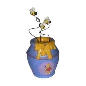 May include: A blue ceramic honey pot ornament featuring a yellow honey drip design at the rim and a small bear character in a red shirt on the front. Three small bees are attached to the top by thin black metal wires extending upwards.