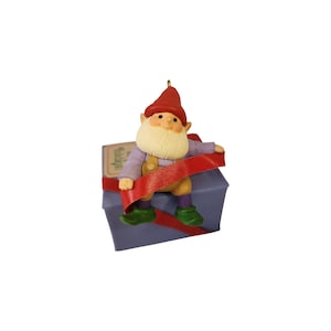 May include: A gnome figurine with a red hat, white beard, light purple long-sleeved shirt, tan overalls, and green shoes. The gnome sits on a light purple square gift box wrapped with a red ribbon, featuring a small gold loop at the top.