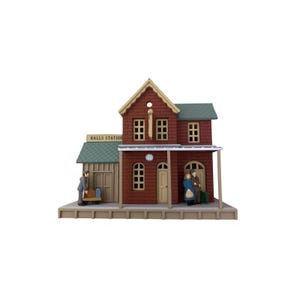 May include: A miniature model of a two-story brick train station named Halls Station. The structure features a green shingled roof, multiple windows, a clock, and a covered platform. Three small figures are positioned on the platform with luggage.