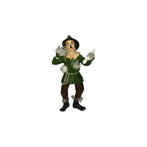 Puede incluir: Una figura de espantapájaros con un sombrero marrón oscuro, una chaqueta verde con detalles de paja y pantalones marrones. La figura tiene la cara pintada, botas negras y sostiene un pergamino blanco enrollado, con paja en las muñecas.