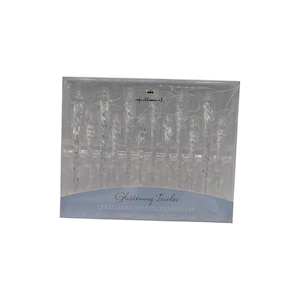 May include: A set of 12 clear, twisted icicle ornaments from the Hallmark Glistening Icicles Frostlight Fairies Collection. The ornaments are packaged in a rectangular clear plastic box with a light blue label featuring the text Glistening Icicles.