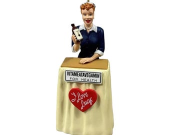 May include: A figurine of a person with red hair wearing a dark blue collared shirt, holding a small brown bottle. The figure stands behind a cream-colored podium with a sign reading VITAMEATAVEGAMIN FOR HEALTH and a red heart shaped I Love Lucy logo.