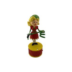 May include: A figurine of a blonde character wearing a green hat and a red dress with scalloped edges. The character holds a green ribbon and stands atop a yellow and red drum-shaped base with a diamond pattern and a small circular hole.