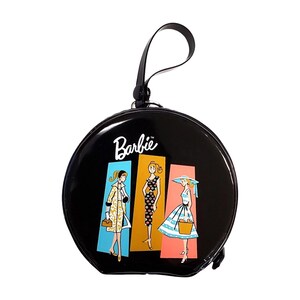 May include: A round black handbag with a wrist strap featuring the white Barbie logo and a graphic print of three figures in different outfits set against blue, yellow, and pink rectangular backgrounds on the front of the glossy circular bag.