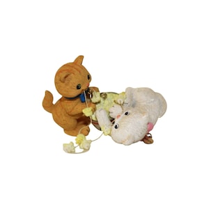 May include: A figurine featuring an orange tabby cat standing and holding a string of yellow yarn that connects to a white cat lying on its side. The white cat is positioned near a small bowl, and both cats have small metallic bells attached to them.