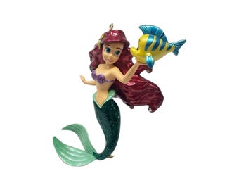 Peut inclure: Une décoration suspendue représentant une sirène aux longs cheveux rouges, portant un haut en coquillages violets et une queue vert foncé scintillante. La sirène tient un petit poisson tropical jaune et bleu, avec une boucle dorée pour suspendre.