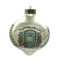 Könnte beinhalten: Ein birnenförmiger, weißer Weihnachtsschmuck mit goldfarbener Kappe. Der Schmuck zeigt eine gemalte Szene einer türkisblauen Tür mit dem Text "1987" und "From Our Home". Der Schmuck hat ein festliches, weihnachtliches Thema.